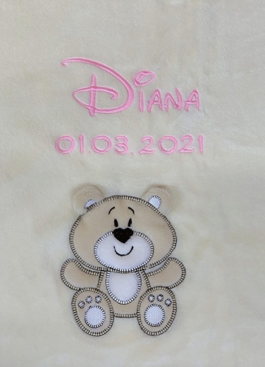 Gros plan sur une couverture pour bébé beige brodée : le prénom « Diana » en lettres cursives roses, avec en dessous la date de naissance « 01/03/2021 ». En bas, une application d’ours en peluche beige souriant.