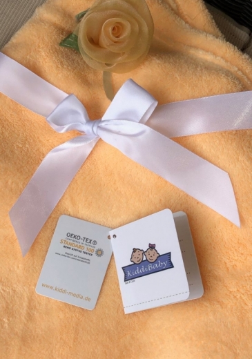 Couverture bébé orange toute douce, avec nœud en satin blanc et rose en tissu jaune clair. Deux étiquettes : Oeko-Tex Standard 100 et KiddiBaby, garantissant sécurité et confort.