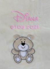 Gros plan sur une couverture pour bébé beige brodée : le prénom « Diana » en lettres cursives roses, avec en dessous la date de naissance « 01/03/2021 ». En bas, une application d’ours en peluche beige souriant.